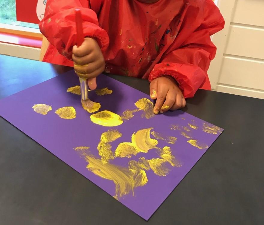 Barn som måler med pensel, gult måling på lilla kartong - Klikk for stort bilete